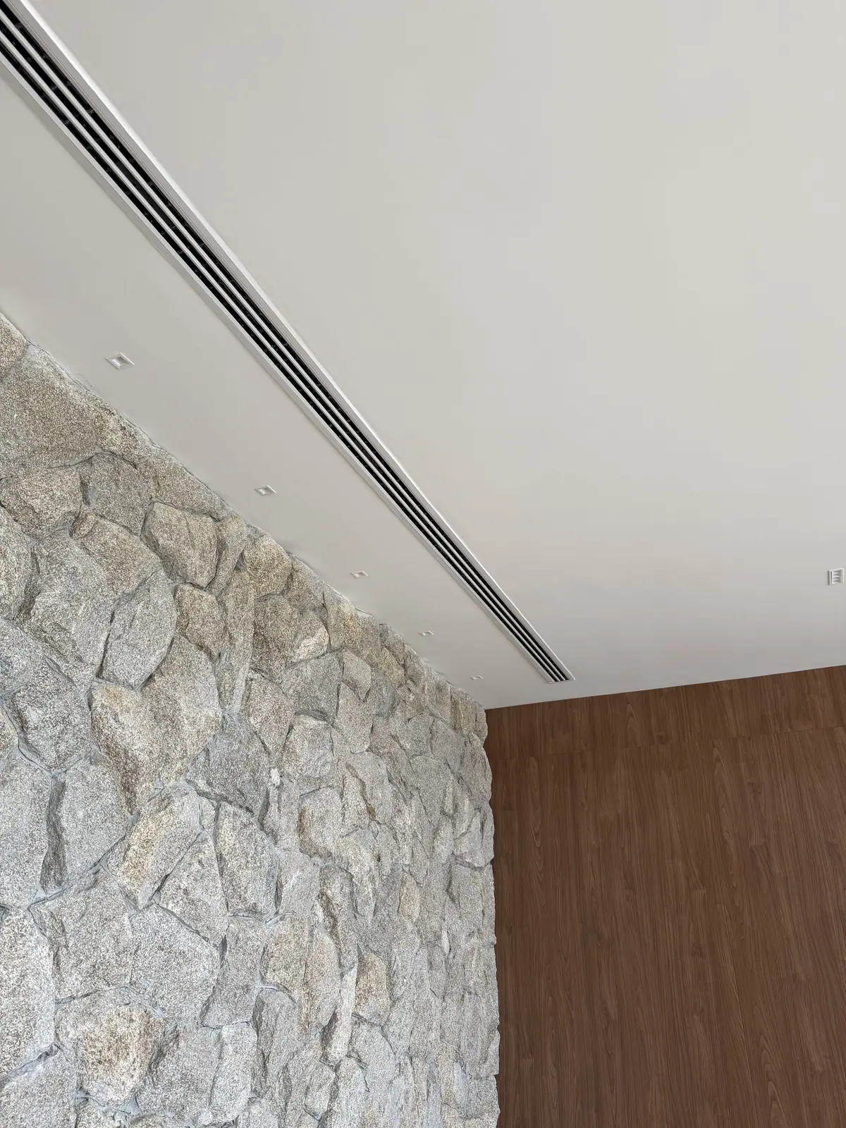 Long linear slot diffuser recessed into a white ceiling beside a stone wall and walnut paneling