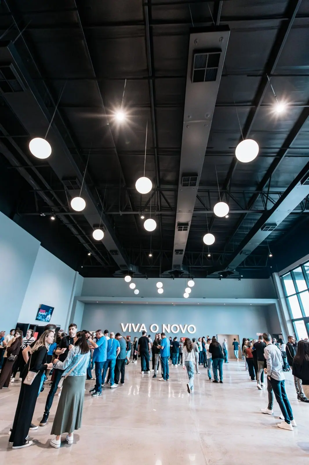 Large open hall with a black exposed ceiling, a long duct run and round pendant lights, with people gathered below