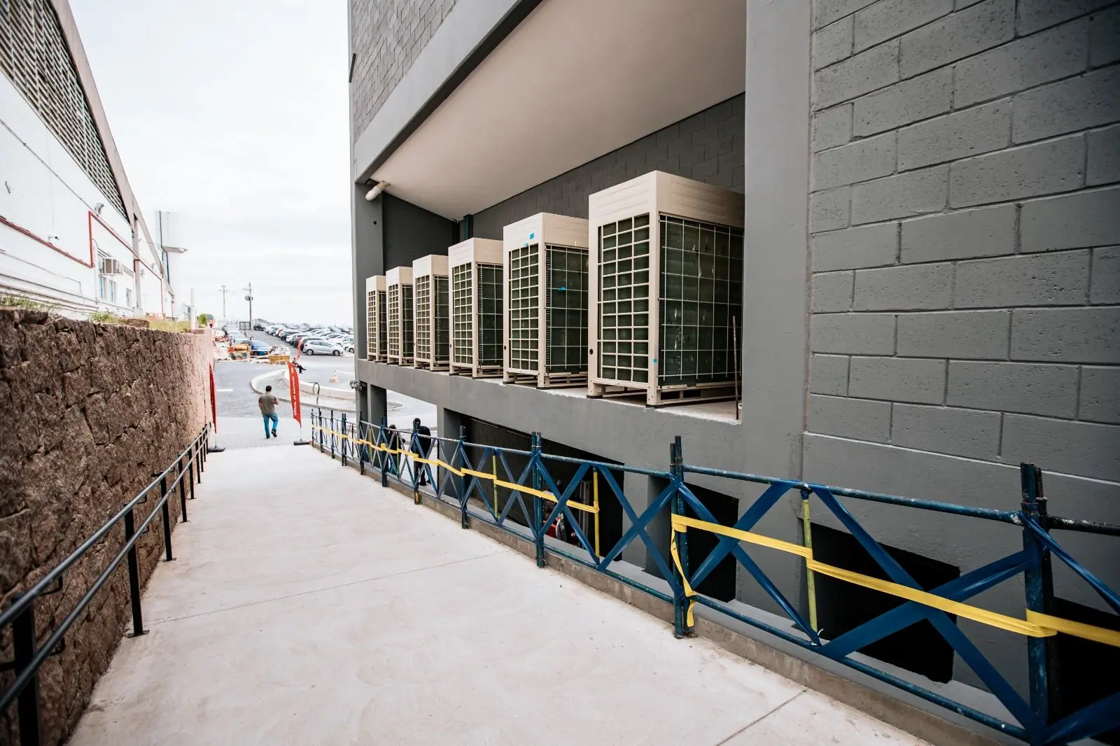Row of six VRF outdoor units on the exterior of a multi-level concrete-block building beside a walkway